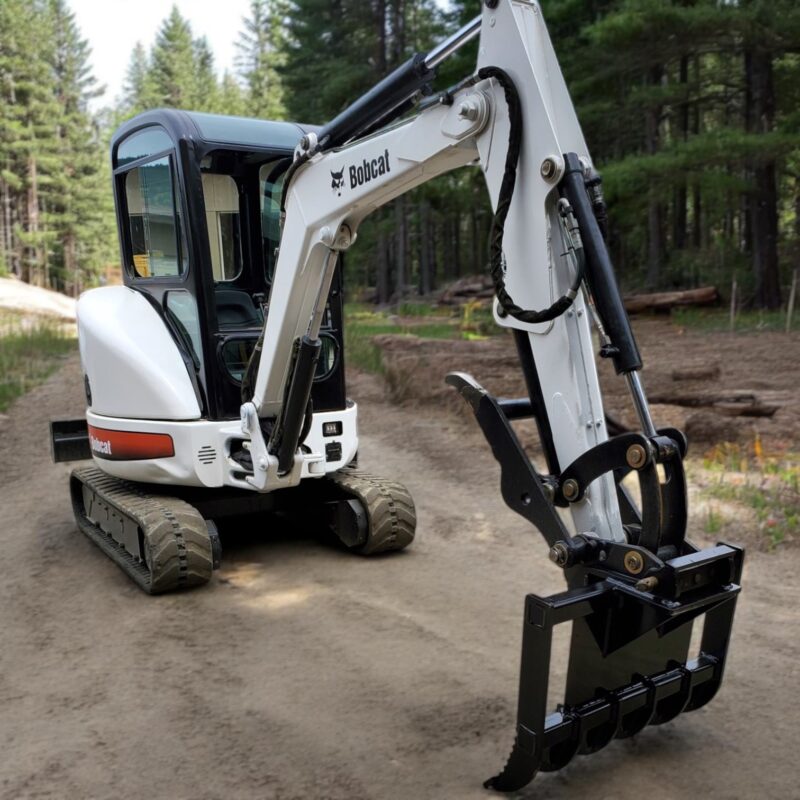 Small Bobcat excavator equipped with brush rake attachment removing brush and vegetation in dense forest